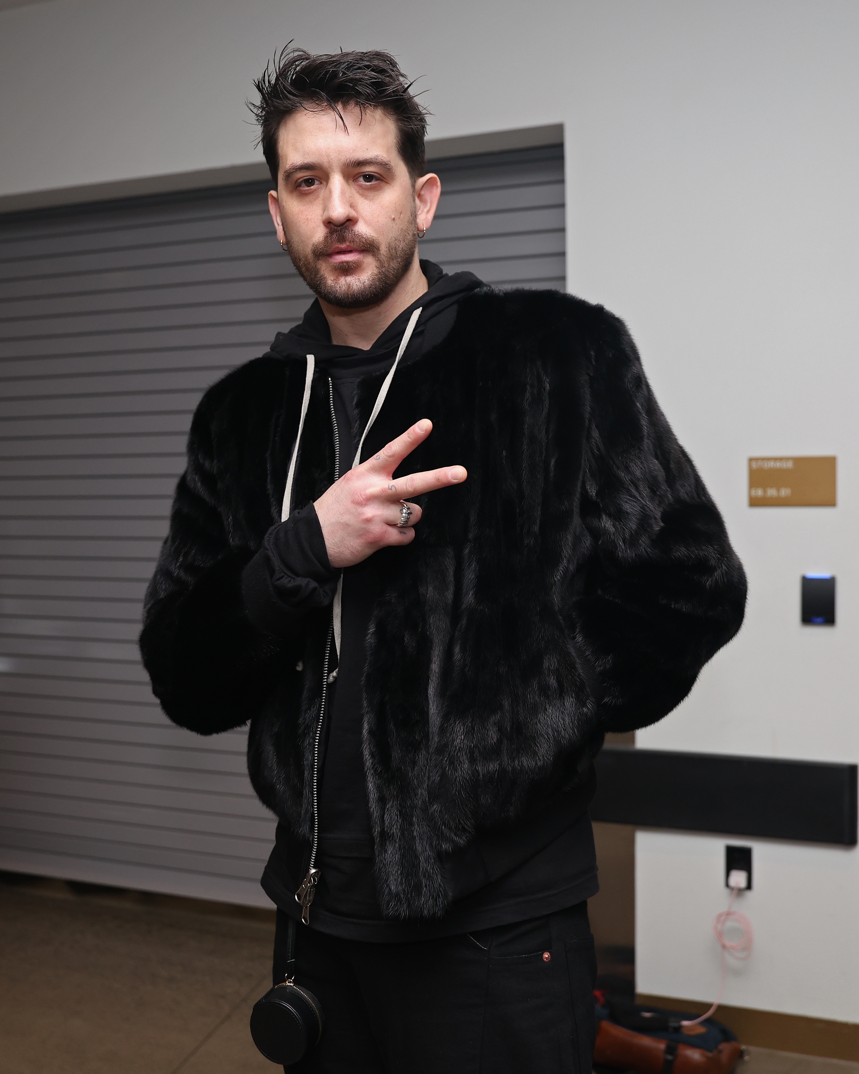 G-Eazy wearing a plush jacket and hoodie makes a peace sign gesture, posing casually at an indoor event