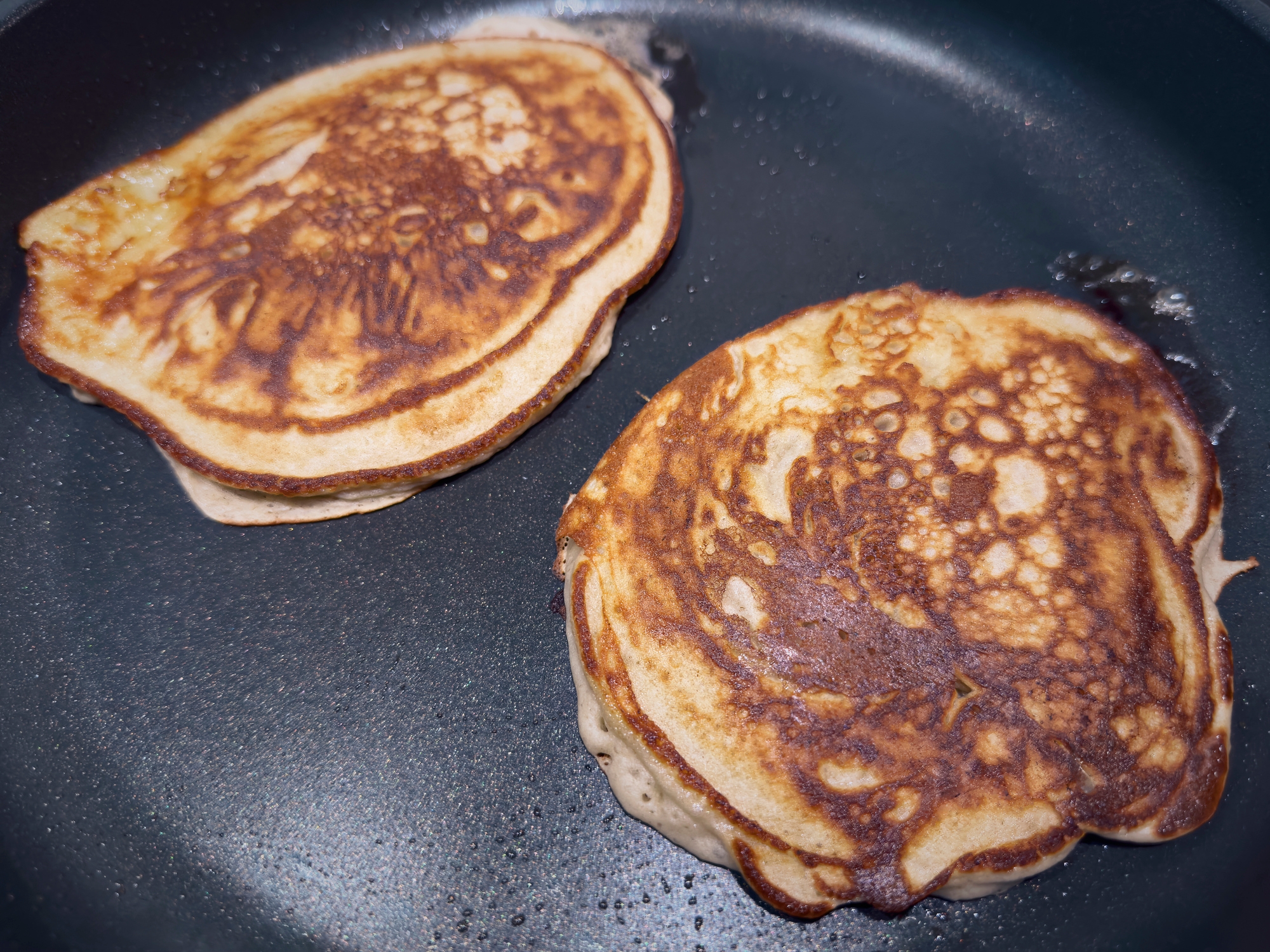Two pancakes cooking in a pan. They are golden brown with a slightly textured surface