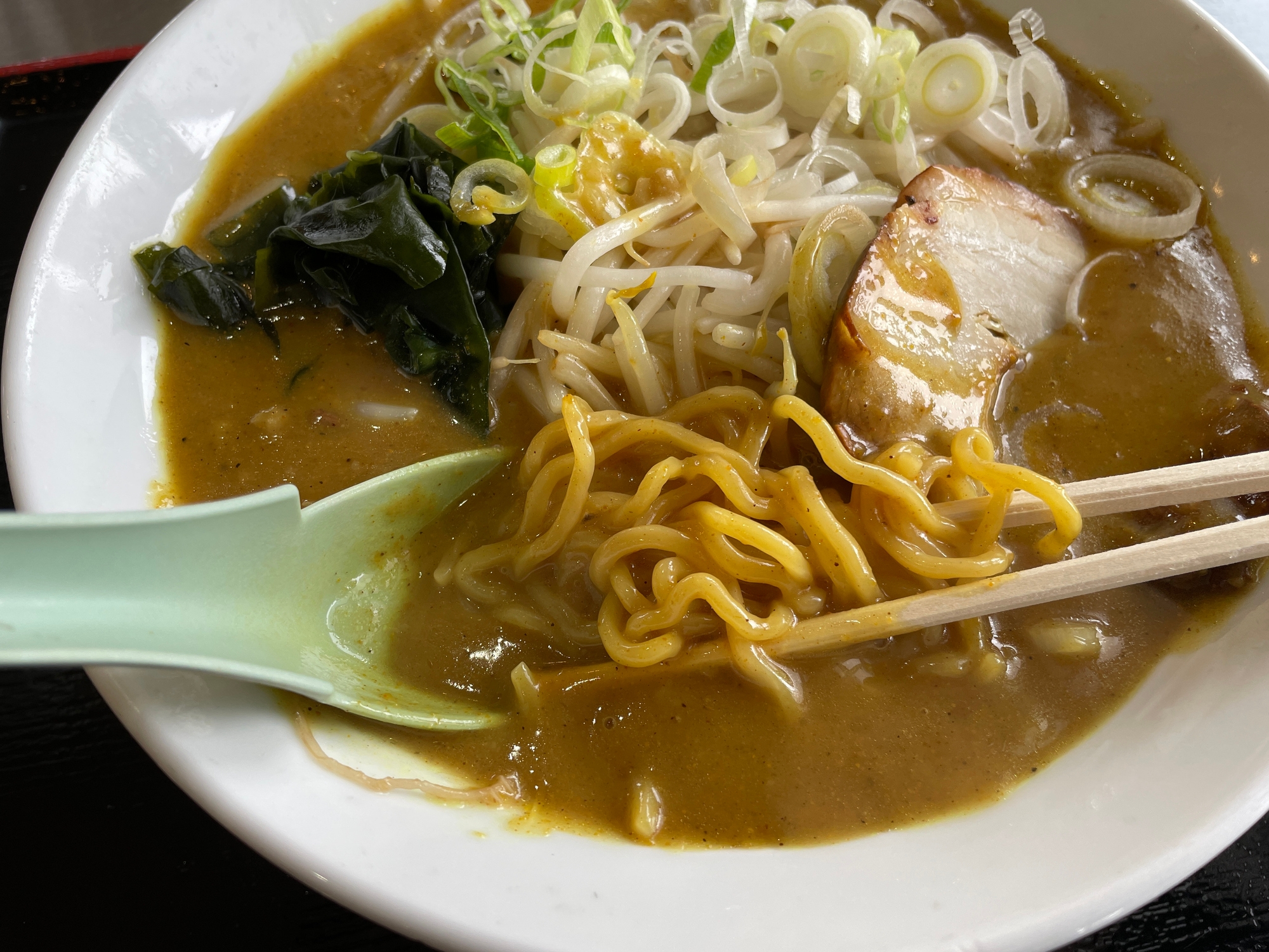 Bowl of ramen with noodles, broth, sliced pork, bean sprouts, seaweed, and green onions, with a spoon and chopsticks