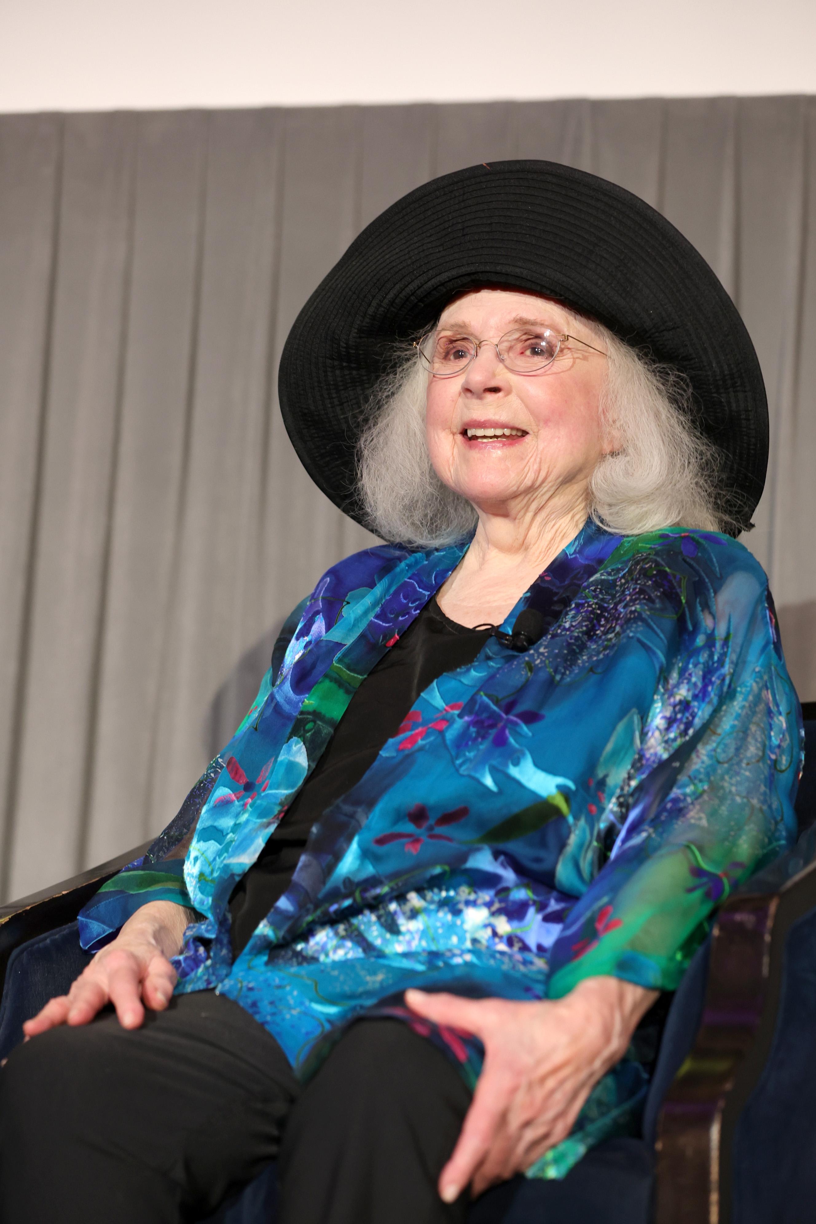 Piper Laurie seated, wearing a wide-brimmed hat and a flowing patterned jacket, smiling during an event