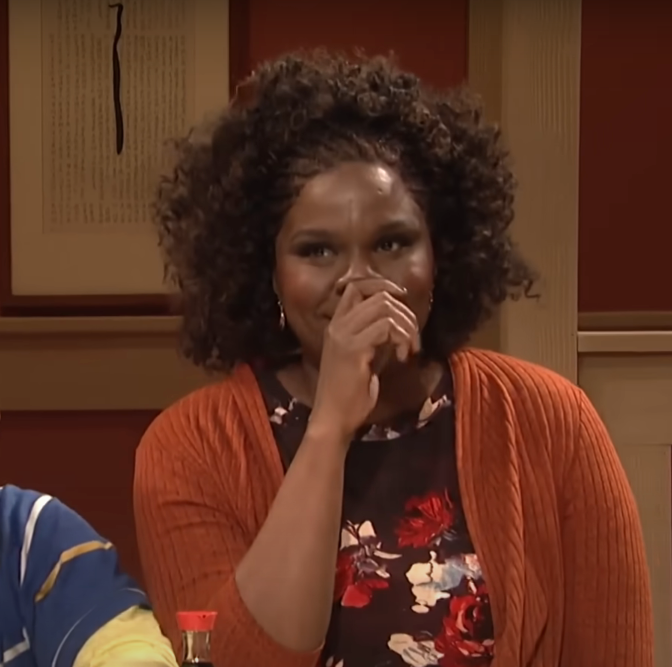 A person with curly hair in a floral blouse and orange cardigan covers their mouth, appearing amused or surprised, in an indoor setting