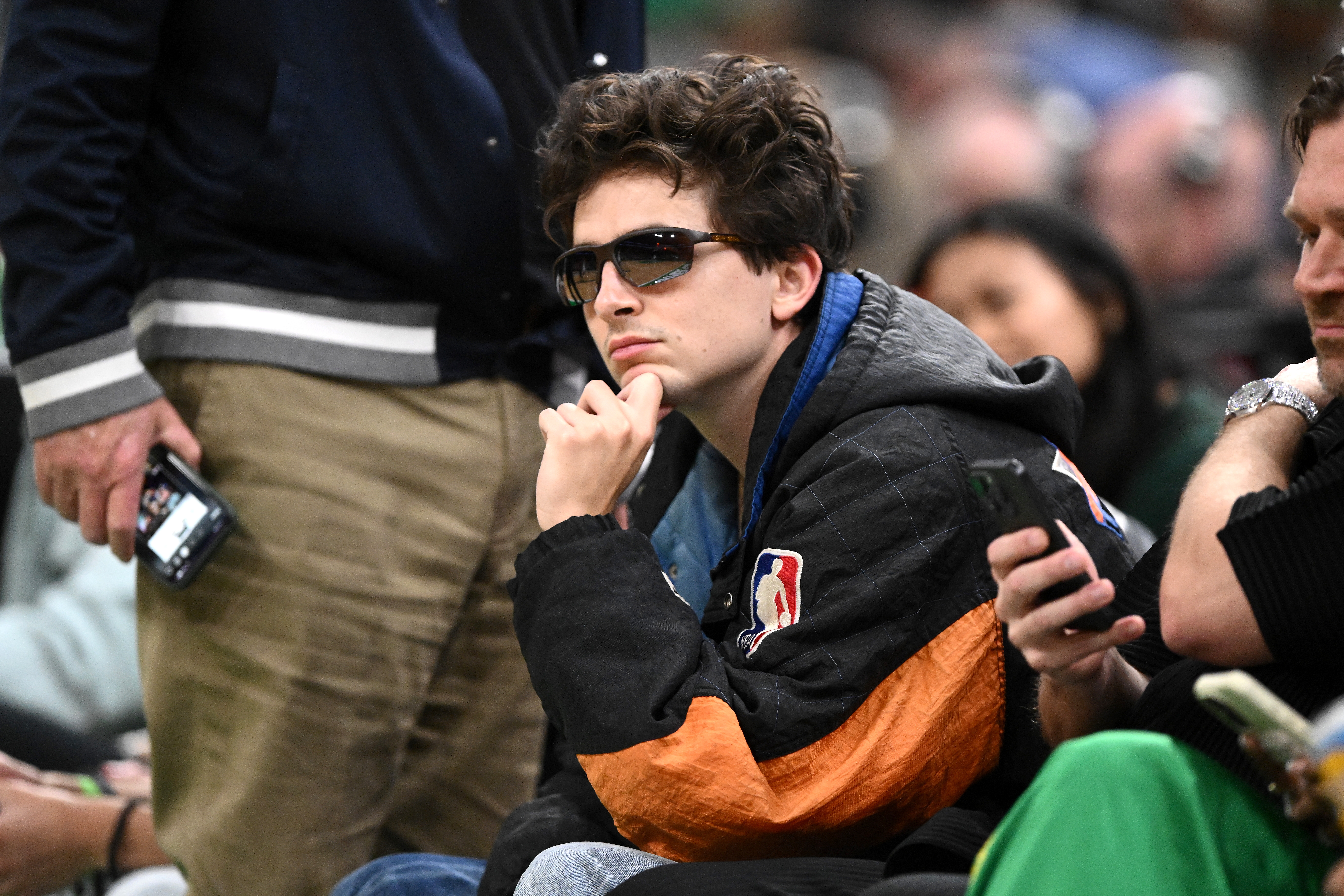 Person in a basketball jacket and sunglasses seated among a crowd, looking thoughtful