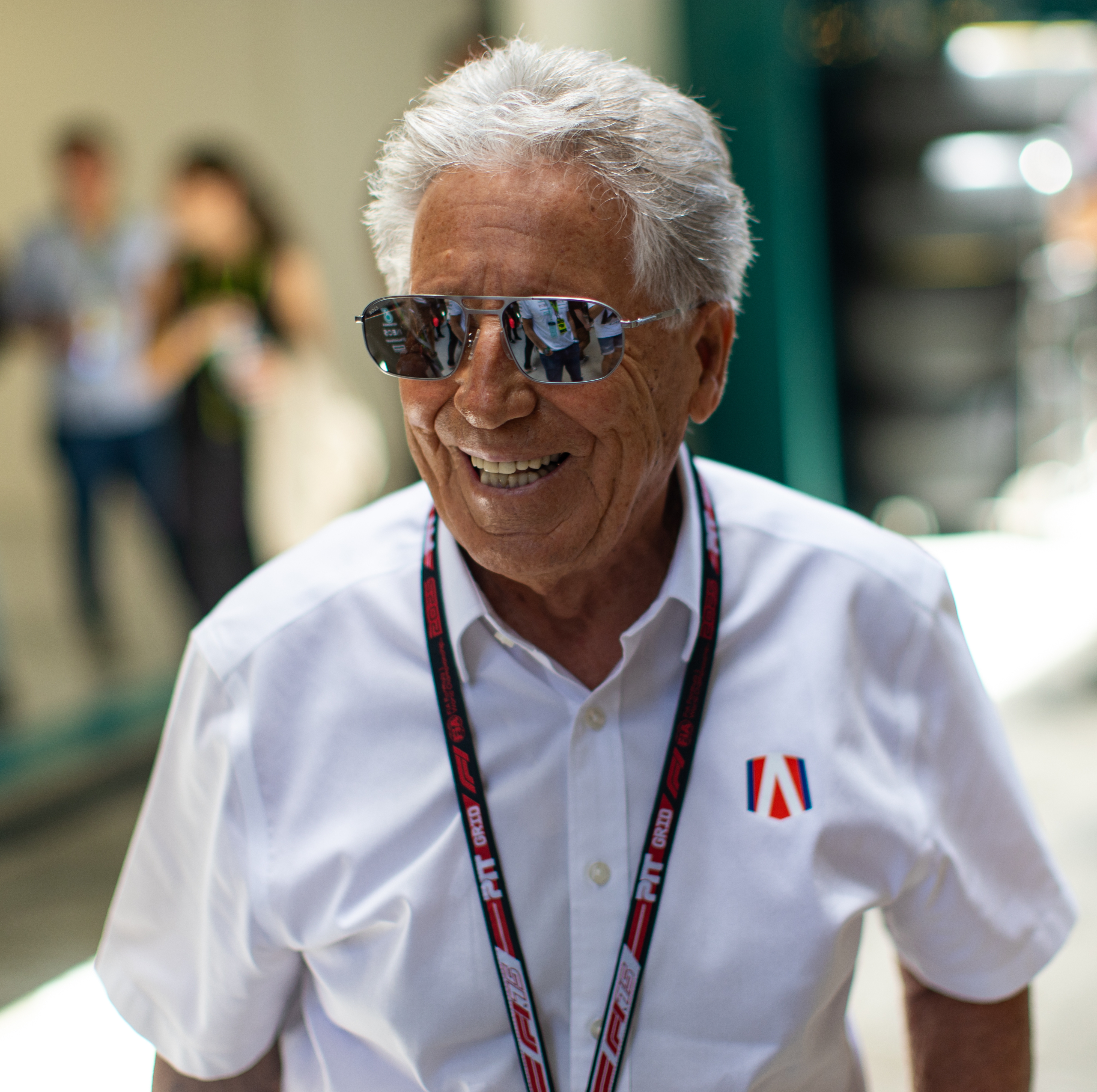 Mario Andretti with sunglasses, white shirt, and a lanyard, smiling outdoors. Others in background are blurred