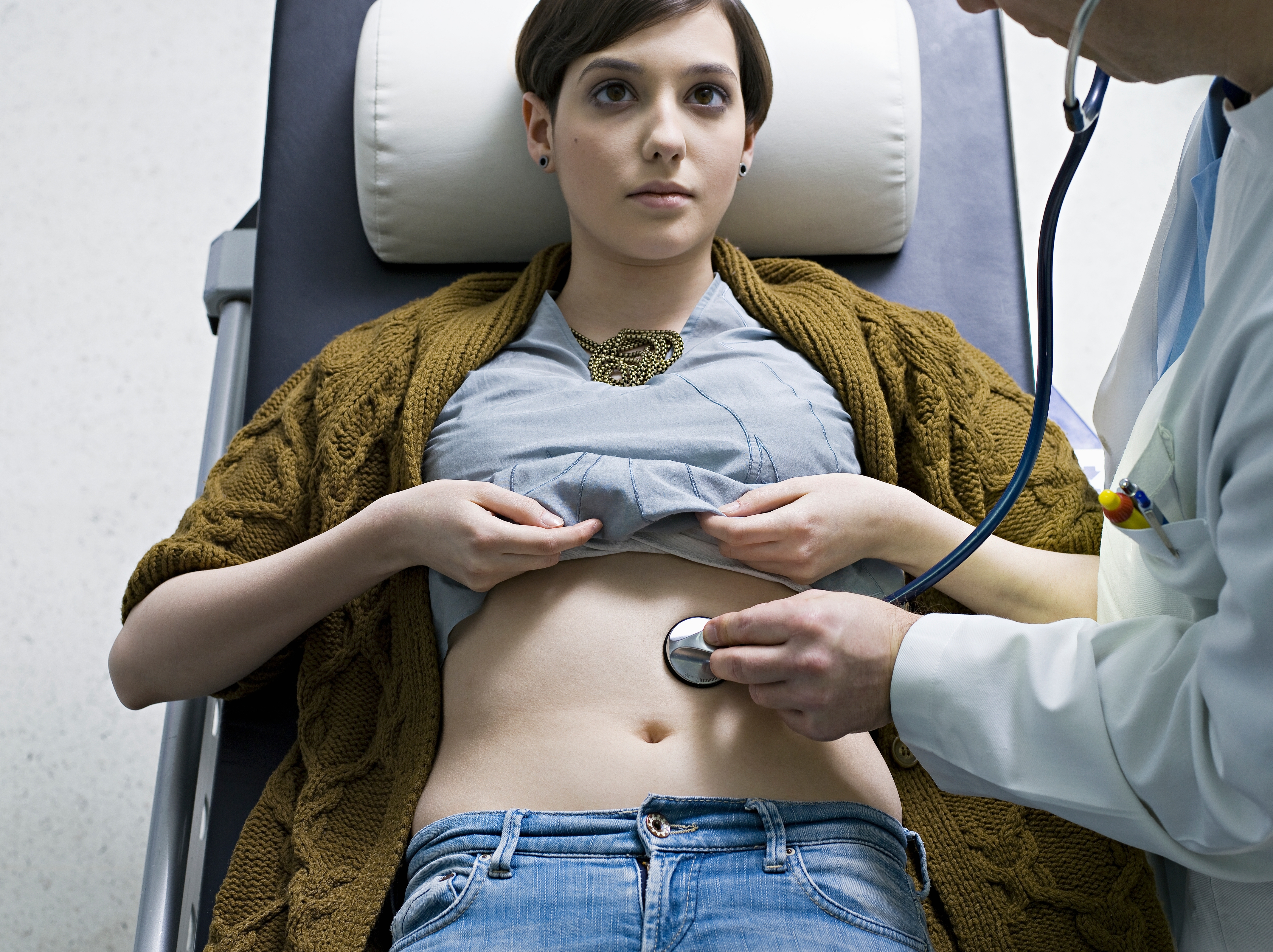 Person lying on exam table, partially lifting shirt for doctor using a stethoscope on abdomen during a medical exam
