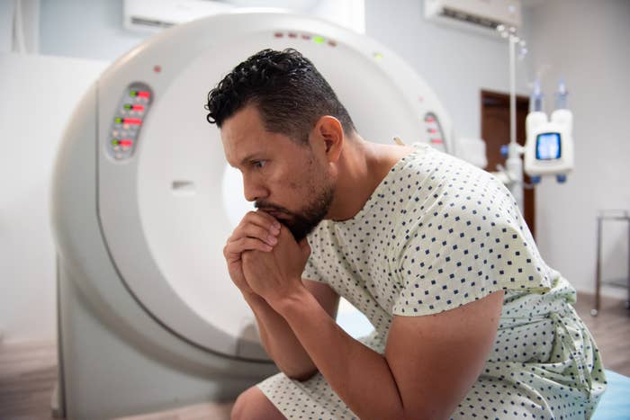 Man in a hospital gown sitting pensively by a CT scan machine, appearing deep in thought