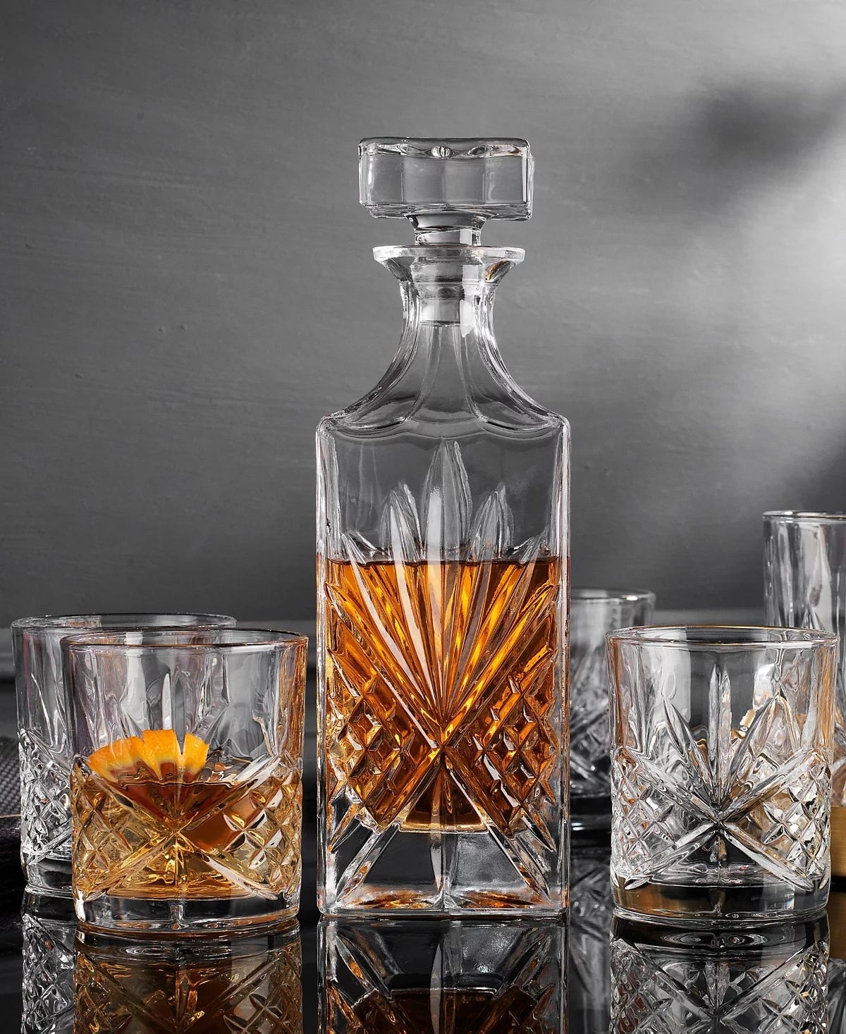 Set kristal decanter dengan wiski dan enam gelas yang cocok, satu dihiasi dengan twist oranye, ditampilkan di permukaan reflektif