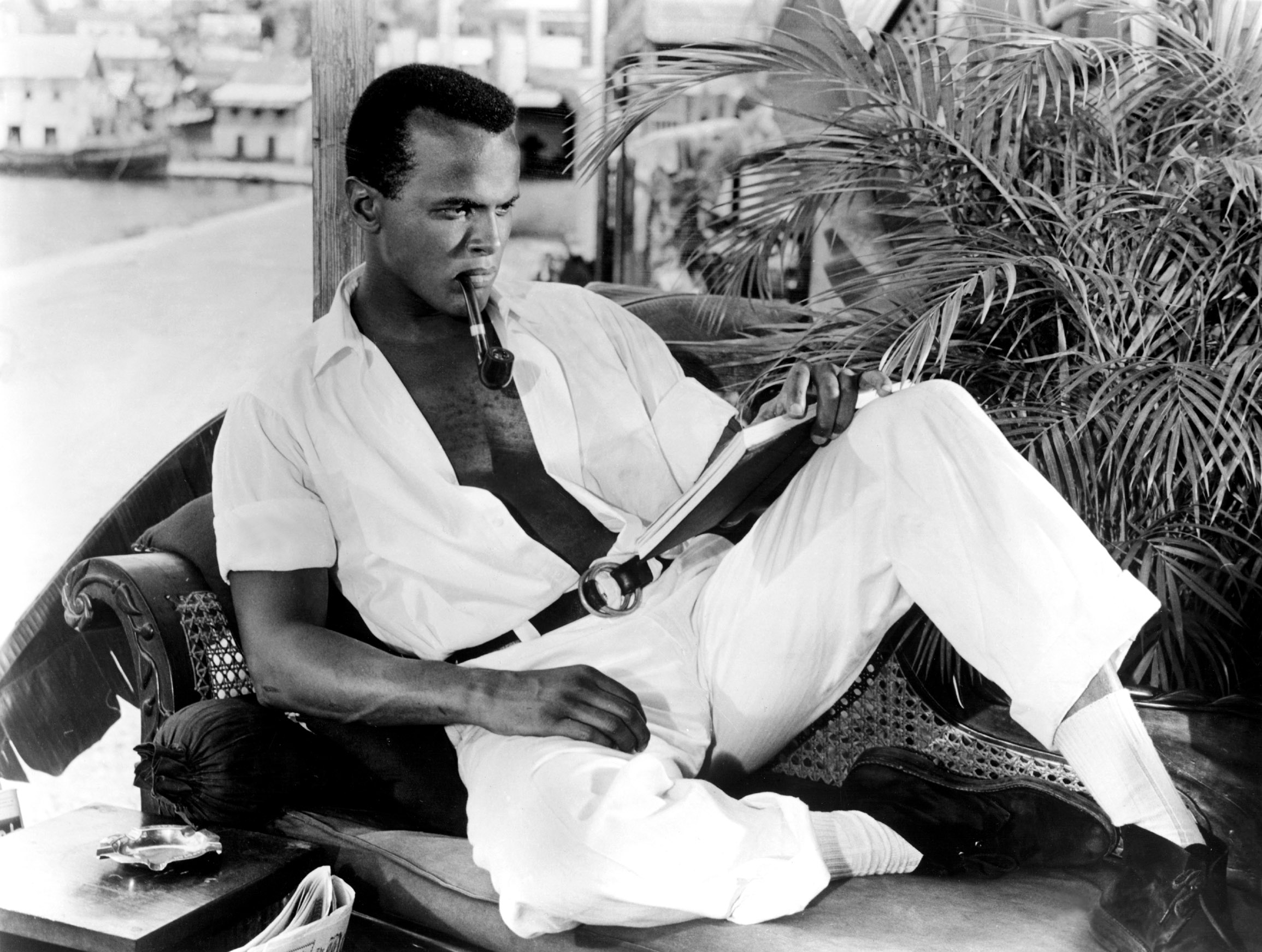 Person lounging on a chair with a cigar, wearing a relaxed open shirt and trousers, reading a book, surrounded by a tropical setting