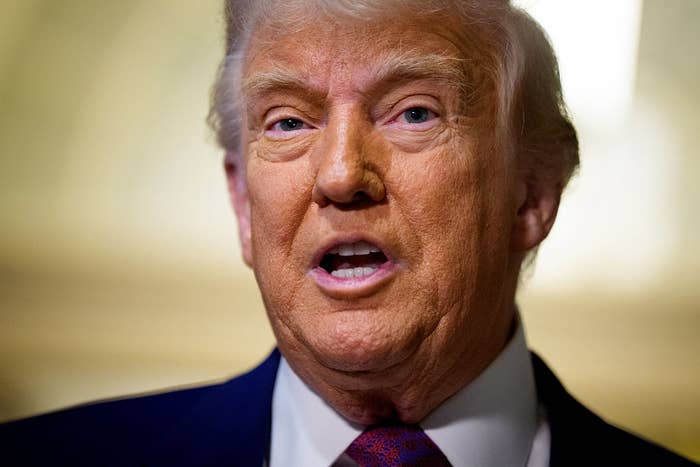 A person speaking, wearing a dark suit and tie. Neutral facial expression