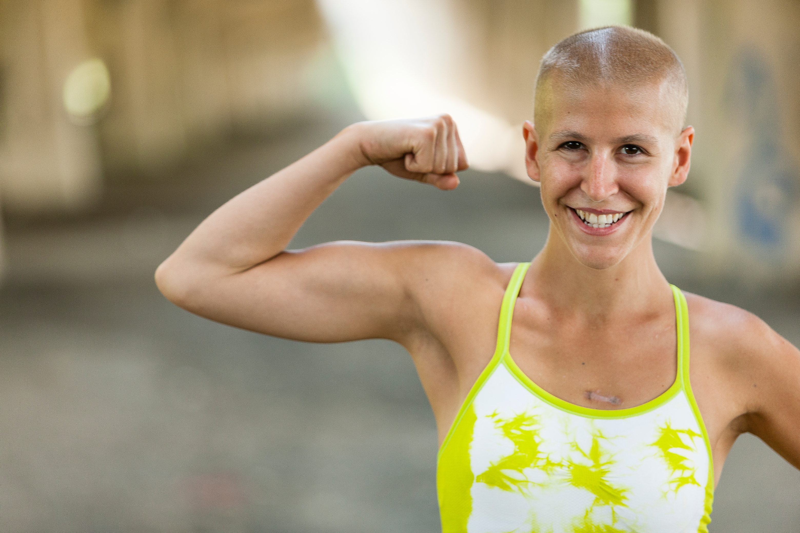 Person with short hair smiling and flexing their arm, wearing a sleeveless top with a vibrant pattern, outdoors in a blurred background
