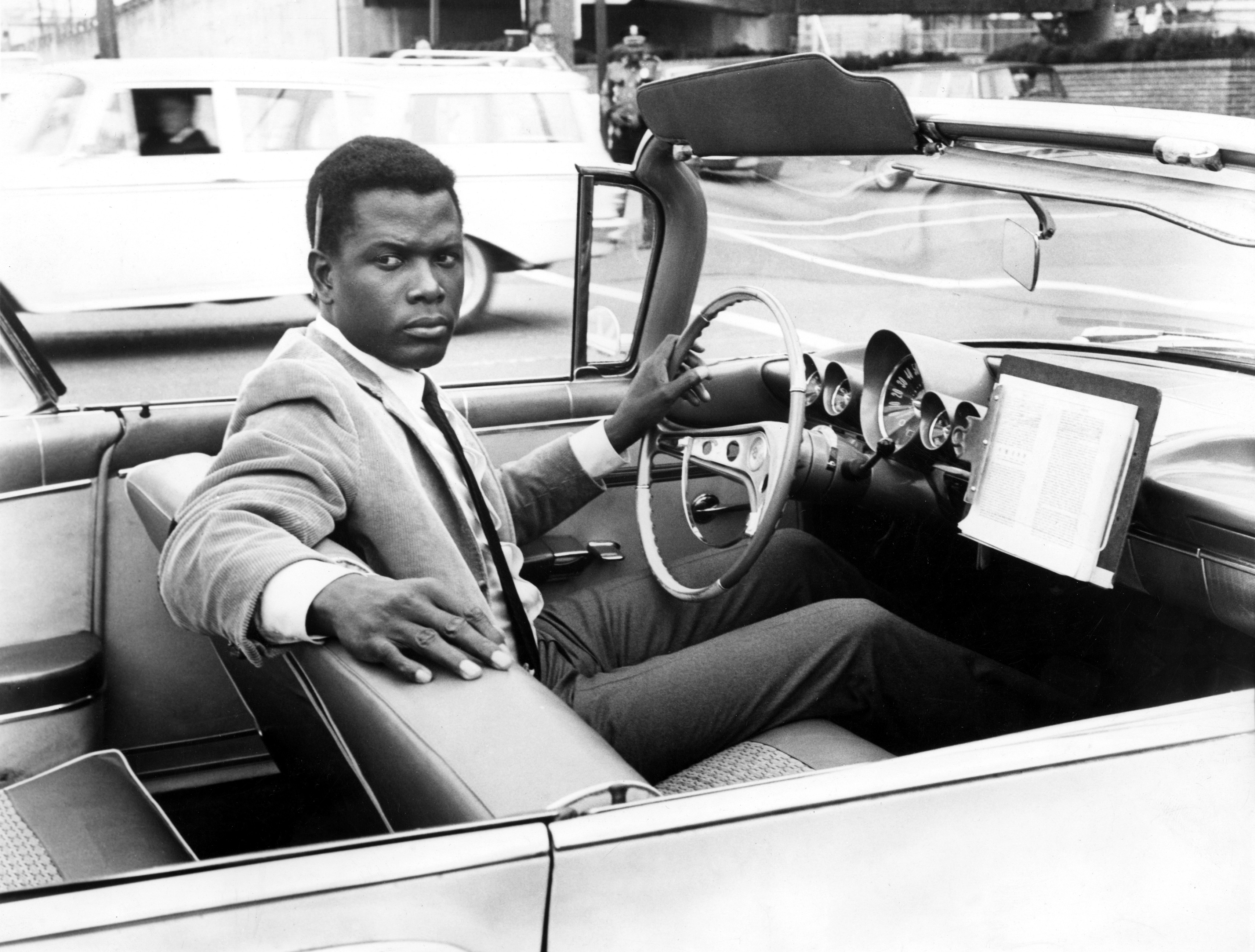 Individual in a suit driving a classic convertible, looking towards the camera, with a city street in the background