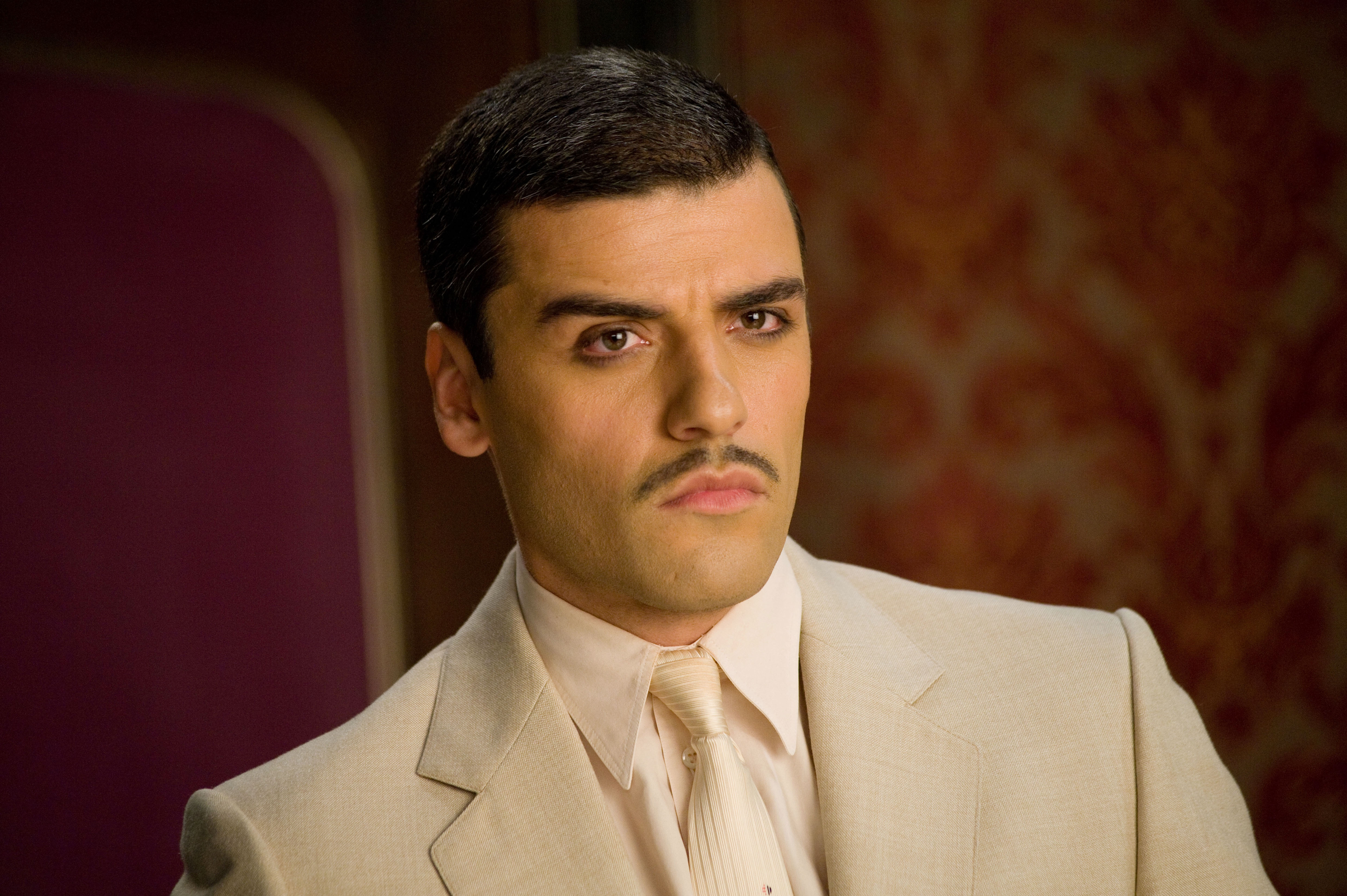 A man with short hair and a mustache wears a classic light suit with a tie, posing in front of an ornate background