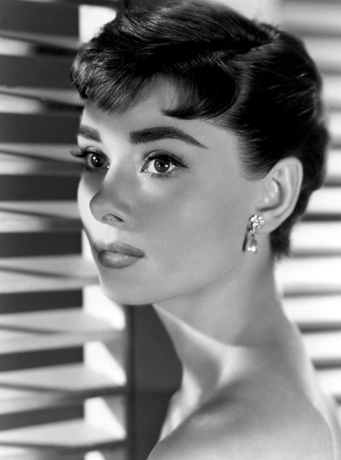 A woman with a classic updo and earrings poses elegantly, gazing to her left, with a patterned backdrop adding depth