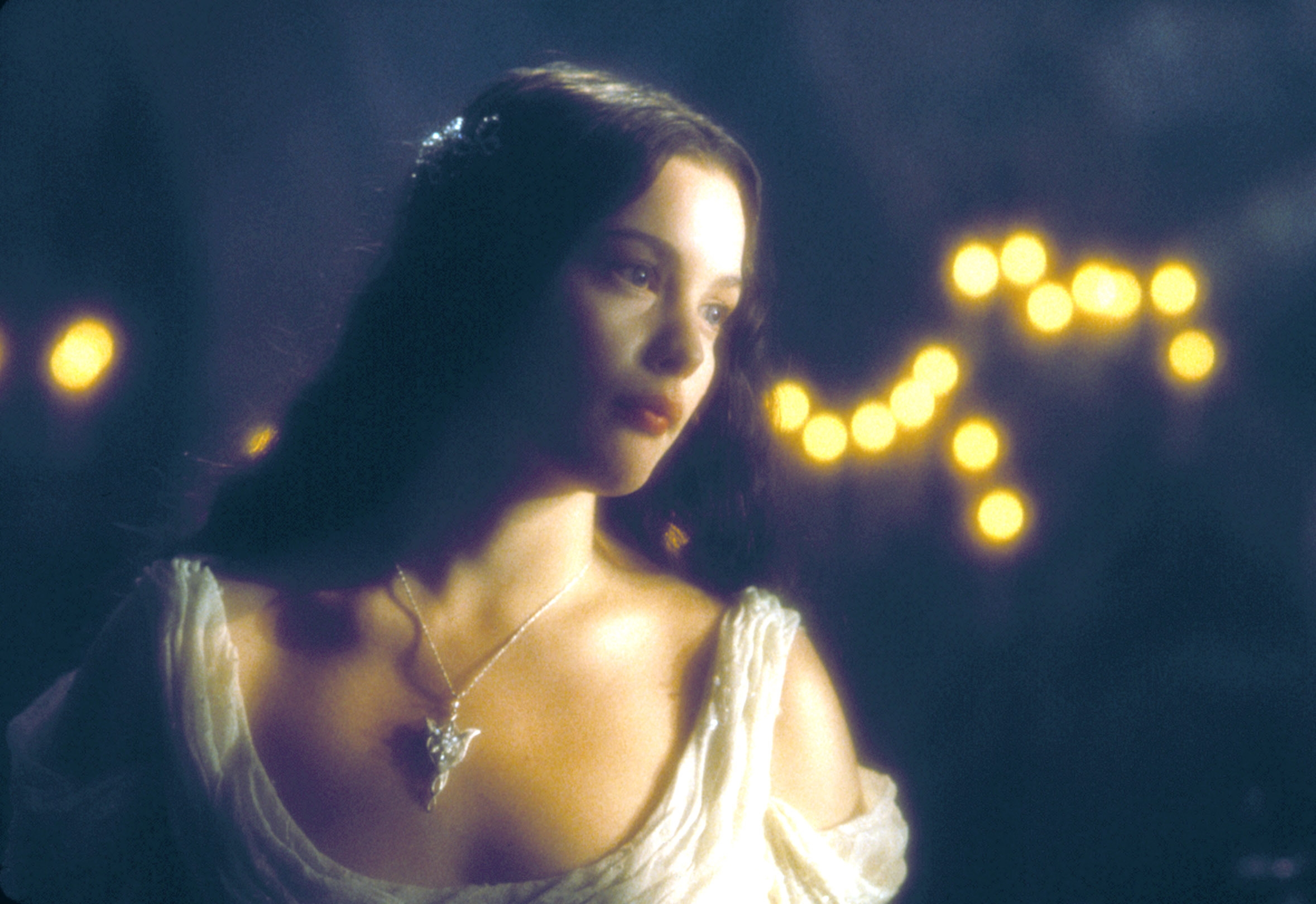 A woman in a flowing gown with a pendant necklace gazes softly, with blurred lights in the background