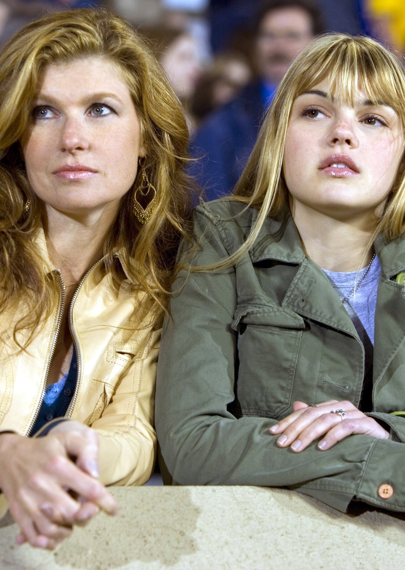 Two women lean on a railing, watching an event. One wears a light jacket, the other a darker one with a pin. They appear focused and engaged