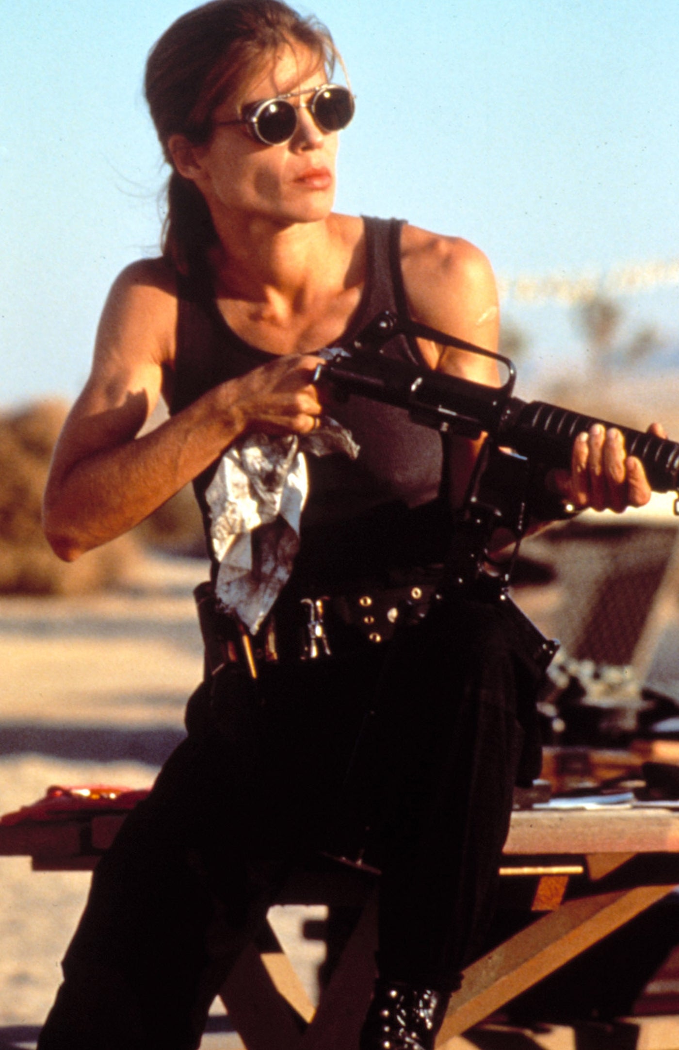 Person in a sleeveless top and sunglasses, sitting on a table, holding a rifle, in an outdoor setting