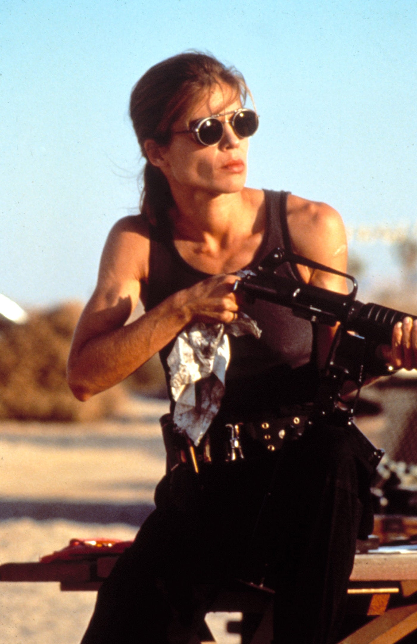 Person in a sleeveless top and sunglasses, sitting on a table, holding a rifle, in an outdoor setting