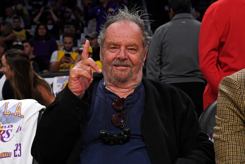 A well-known actor at a basketball game, wearing a casual shirt and jacket, holding up one finger