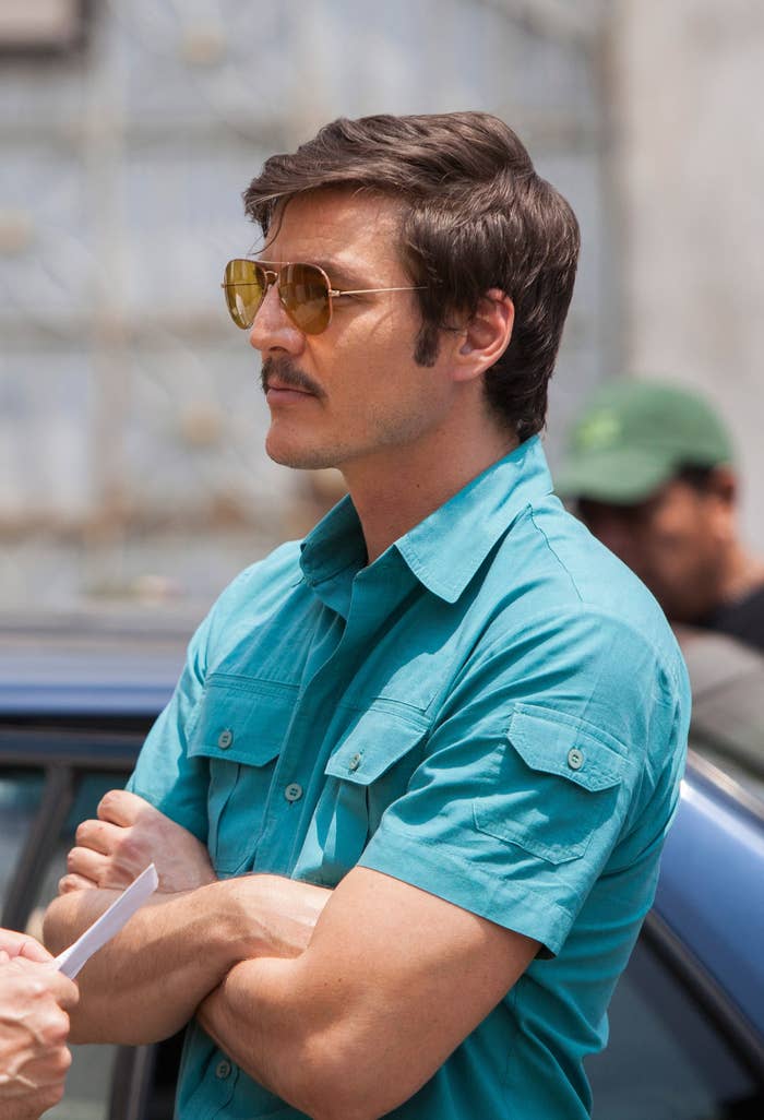 Two men in a casual discussion outdoors; one in a suit, the other in a short-sleeve shirt with sunglasses, arms crossed