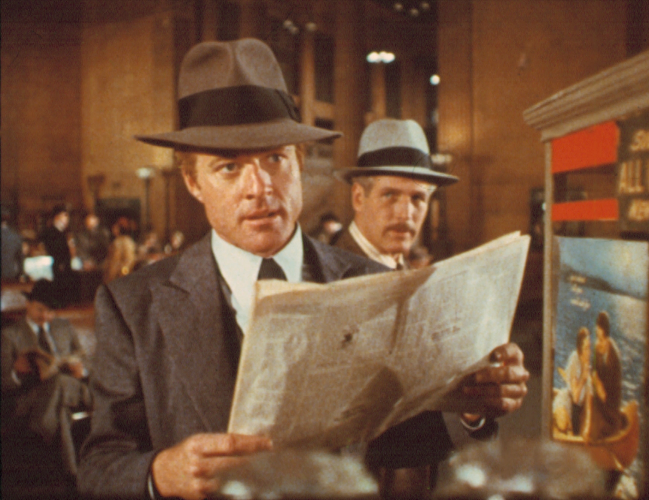Two men in vintage suits and hats read a newspaper in a classic train station setting
