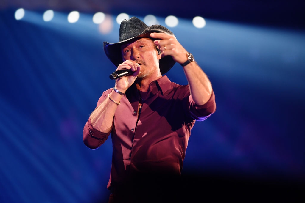 A person on stage performs with a microphone, wearing a cowboy hat and casual shirt, gesturing mid-song during a concert