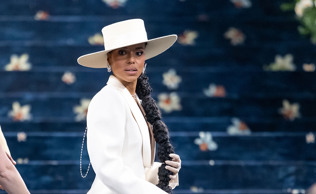 A person on the red carpet wears a stylish white suit with a wide-brimmed hat, holding a braided accessory, against a floral backdrop