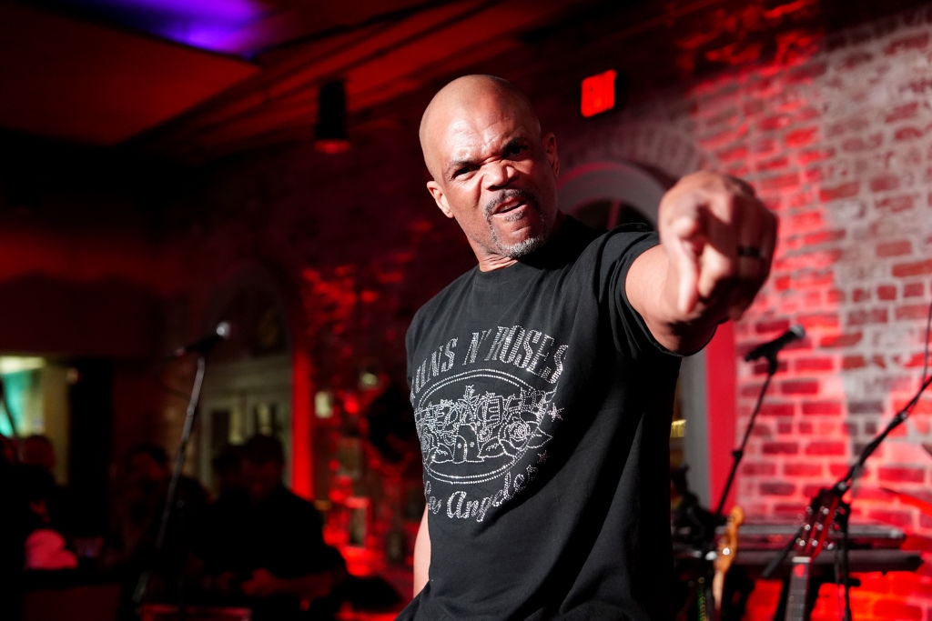A man on stage points assertively while wearing a graphic t-shirt, likely during a performance or event. Mood is intense and dynamic