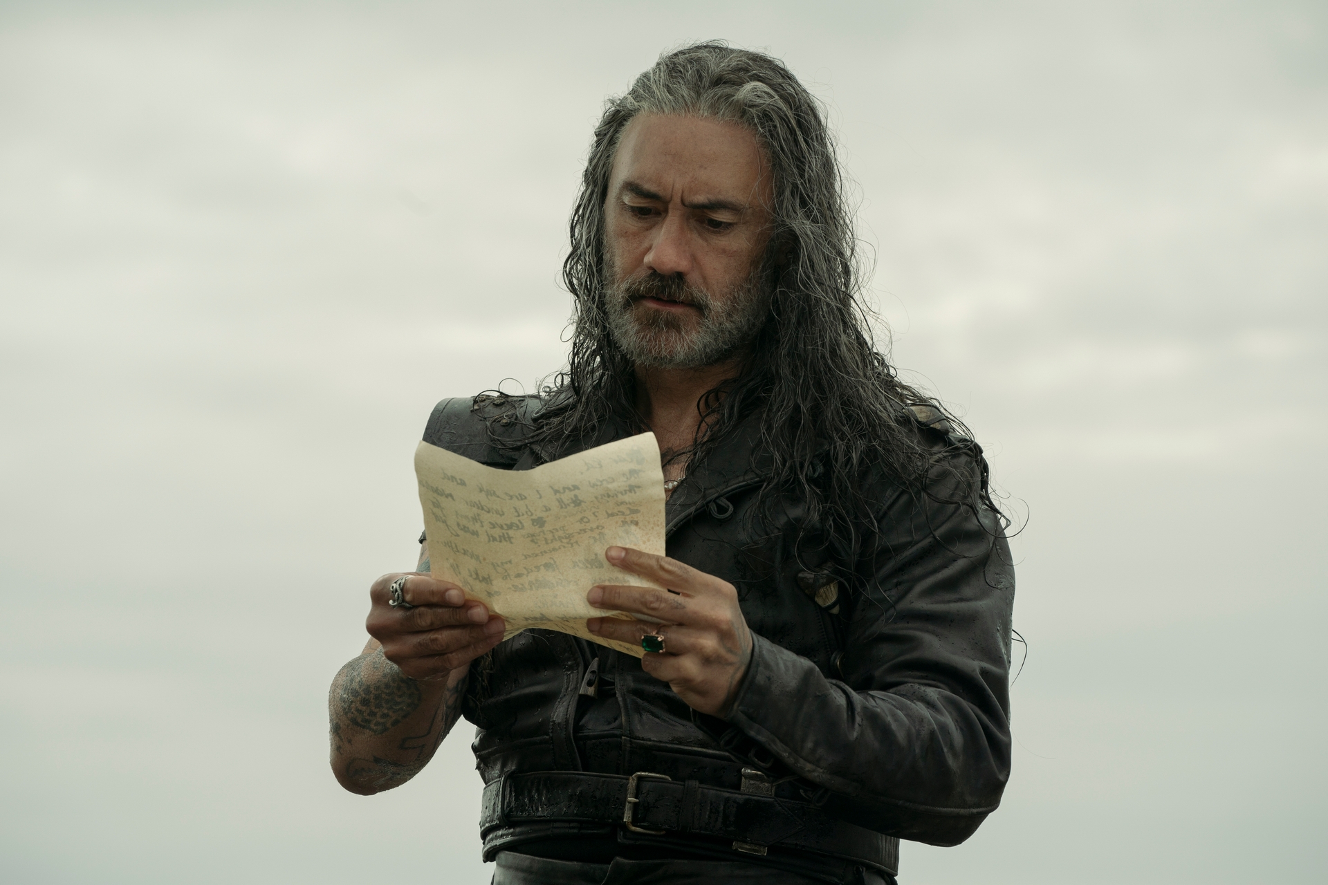 Man with long hair in a leather outfit reads a letter outdoors, looking thoughtful and focused