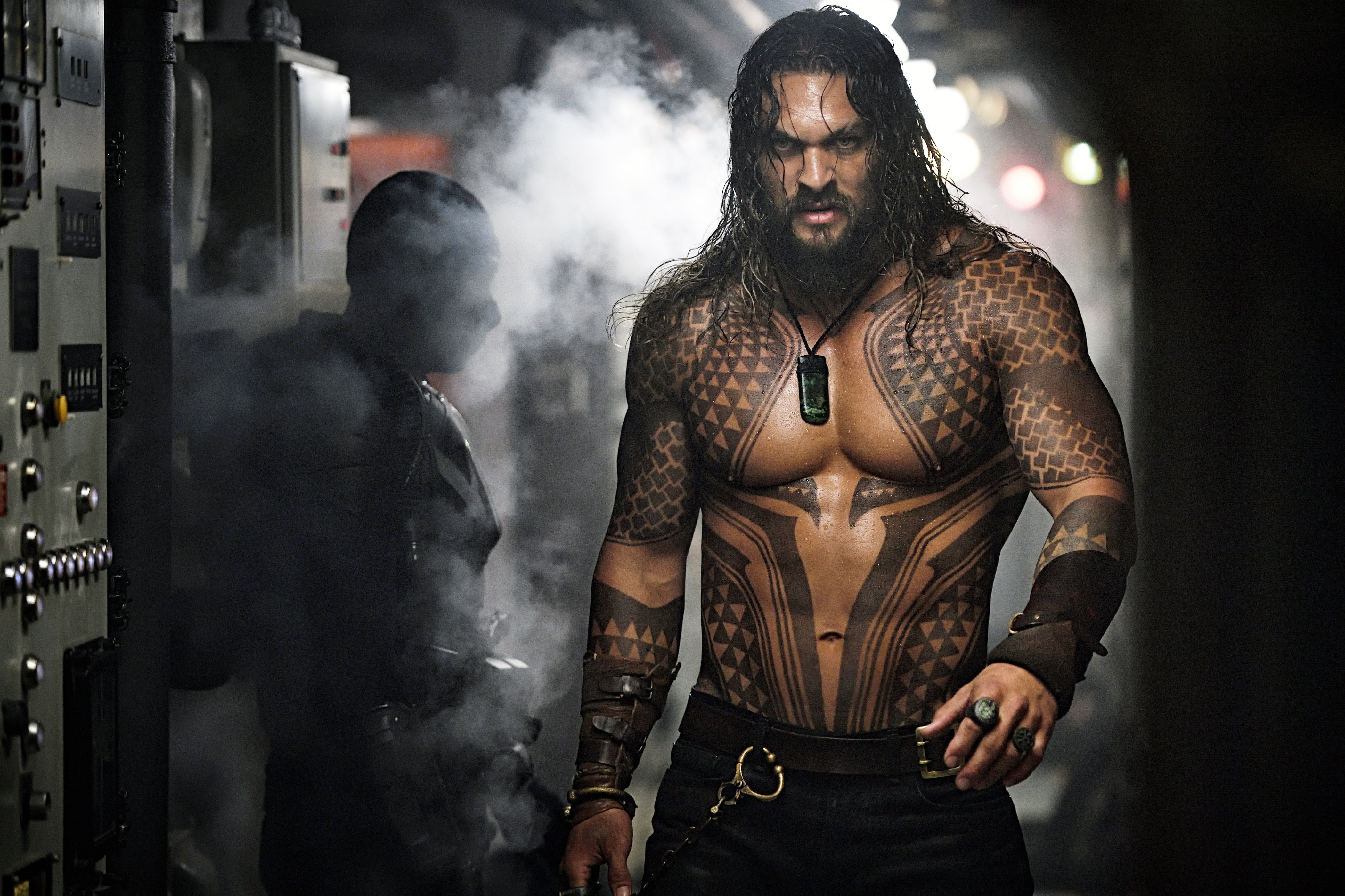 Person with intricate tattoos and long hair walks through a smoky industrial setting, evoking a dramatic and intense atmosphere