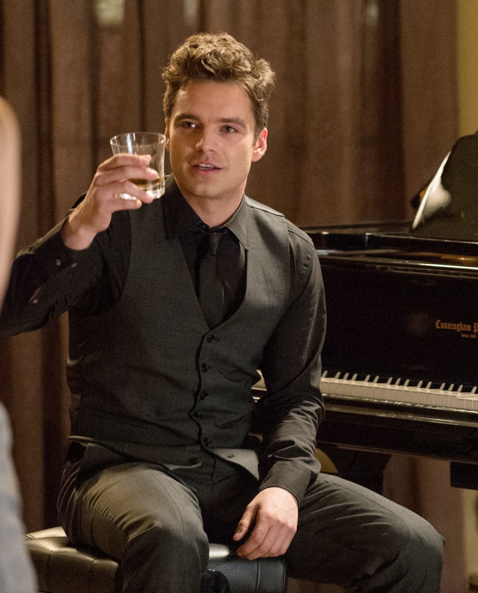 Person in formal attire holding a drink while sitting by a grand piano, engaging with others in a warm, social setting