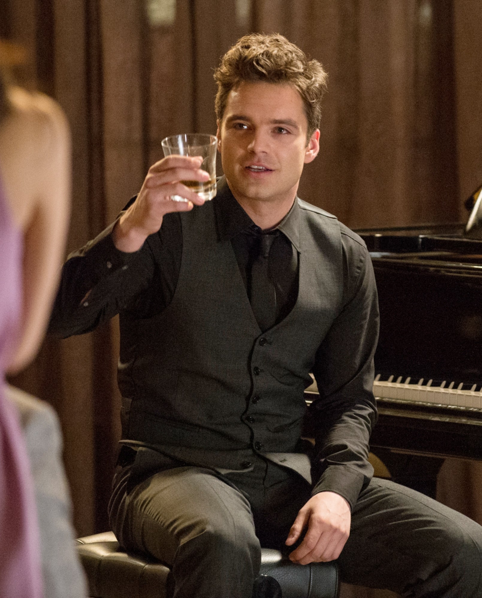 Person in formal attire holding a drink while sitting by a grand piano, engaging with others in a warm, social setting