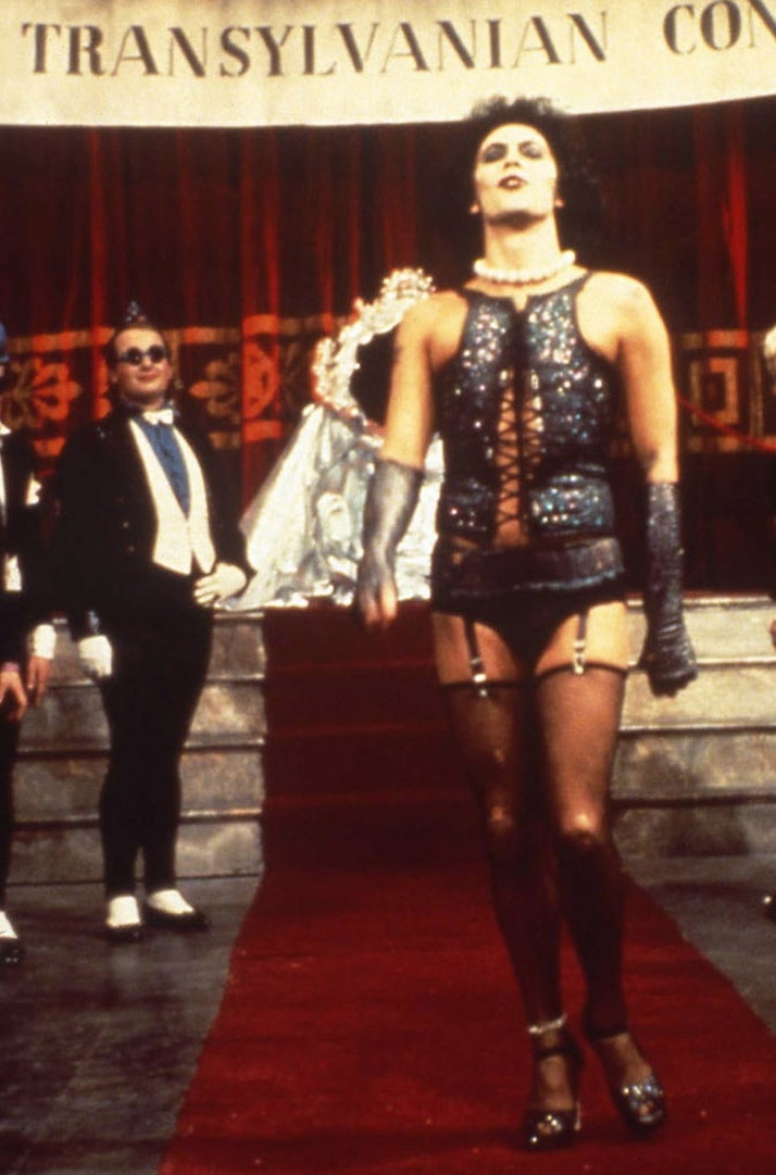 Person dressed in glam rock style corset and fishnets on stage, surrounded by costumed performers at an &quot;Annual Transylvanian Convention.&quot;