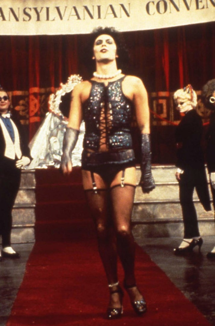 Person dressed in glam rock style corset and fishnets on stage, surrounded by costumed performers at an "Annual Transylvanian Convention."