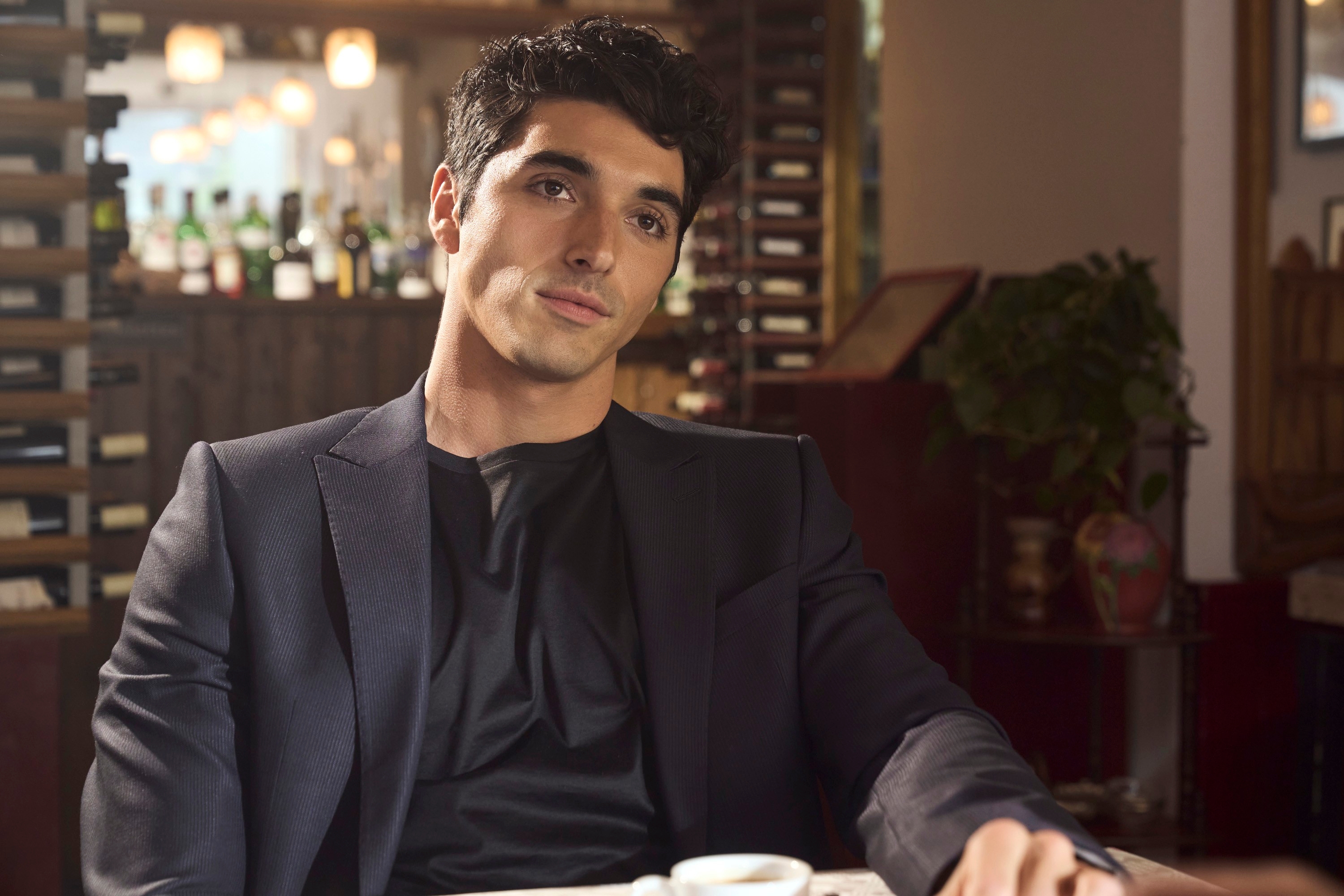 Man in a stylish suit seated at a cafe table with a thoughtful expression