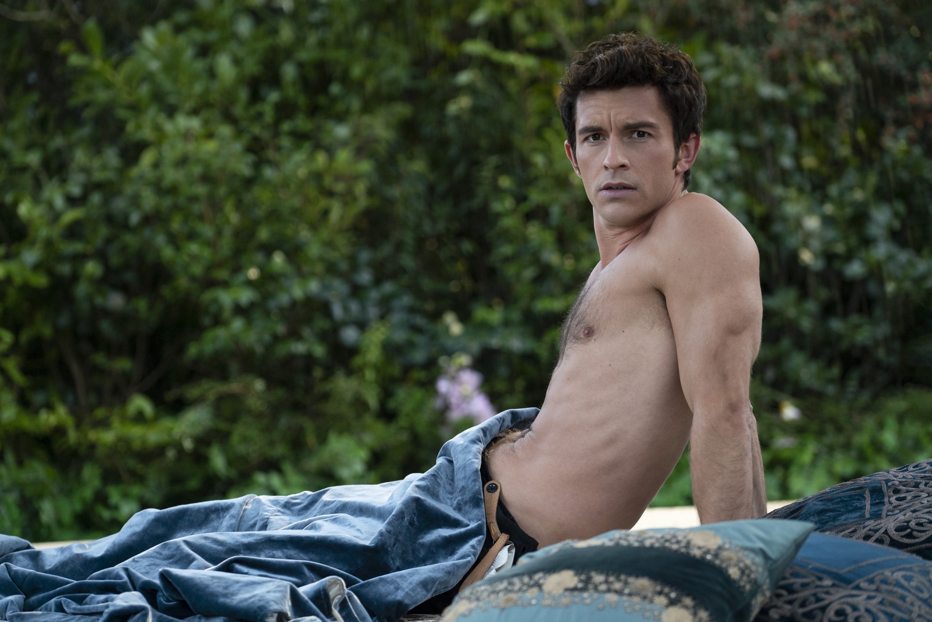 Shirtless man with short hair lying on a cushioned outdoor bench, looking away from the camera with a thoughtful expression