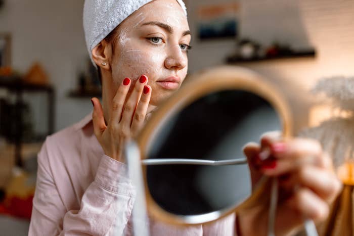 Person applying skincare product, wearing a head wrap, with focus on their reflection in a handheld mirror