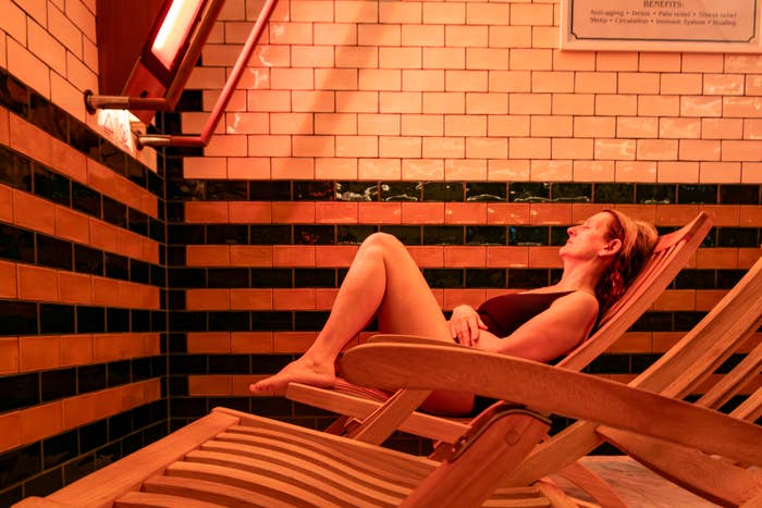 Person relaxing on a wooden lounge chair under a red light in a room with brick walls. They appear calm and at ease