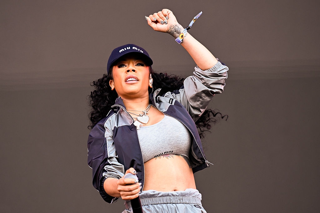 Singer on stage, wearing a cap, cropped top, and track jacket, holding a microphone, arm raised