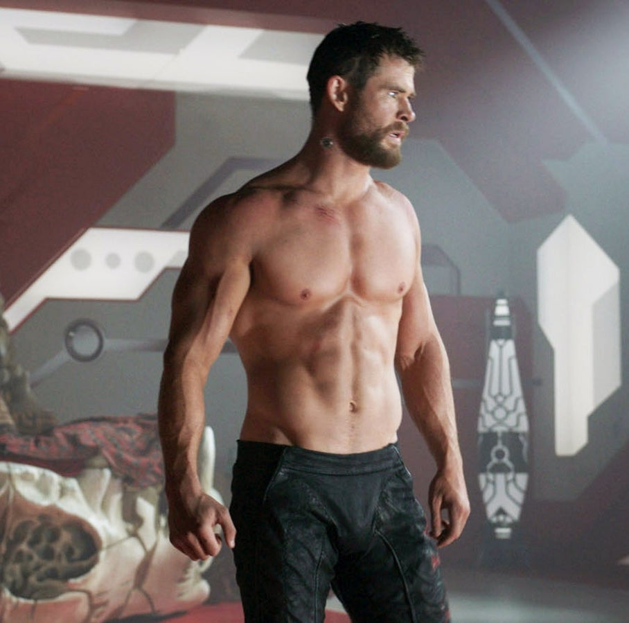 A shirtless man with short hair and muscular build stands in a futuristic setting, looking serious