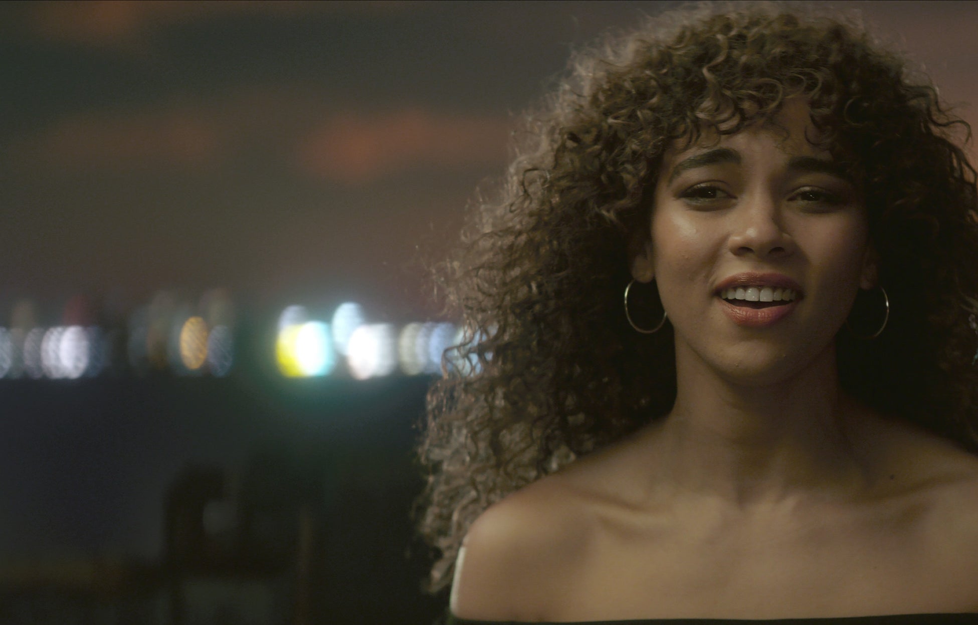 Person with curly hair smiling, standing outdoors at night with city lights blurred in the background