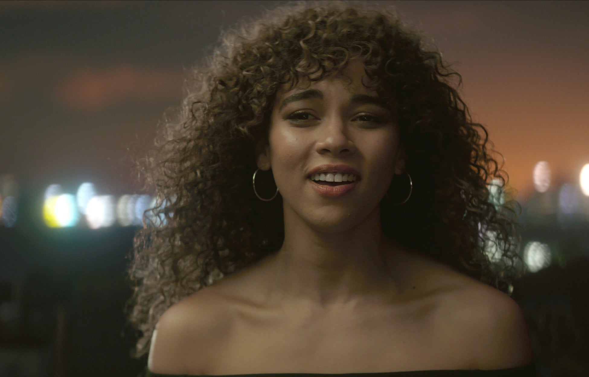 Person with curly hair smiling, standing outdoors at night with city lights blurred in the background