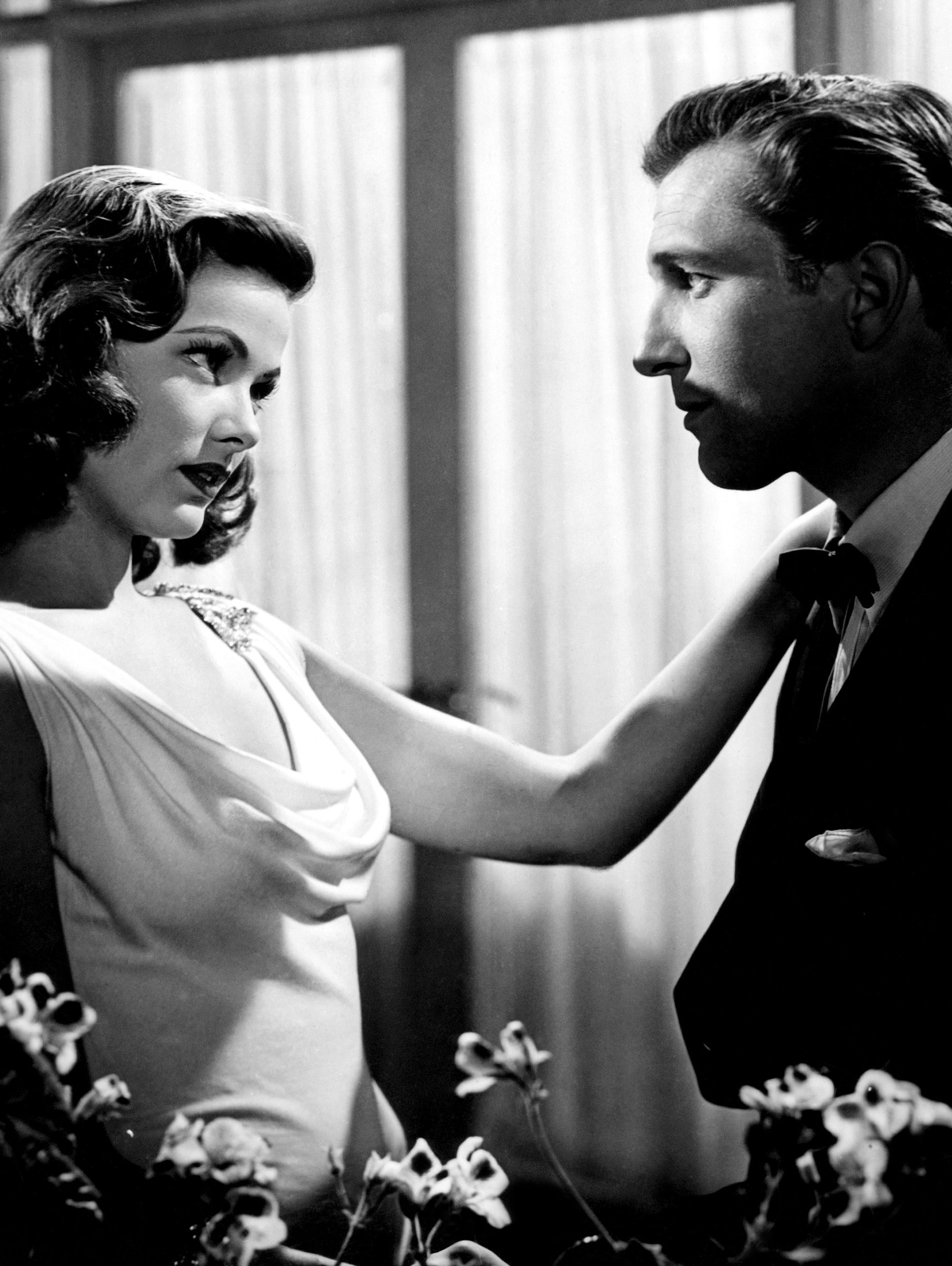 A classic Hollywood scene with a woman in an elegant gown and a man in a suit, gazing intently at each other amidst floral decor