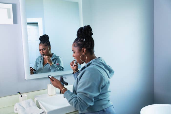 Person brushing teeth while looking at phone in a bathroom. They’re wearing a casual hoodie and earrings, standing at the sink