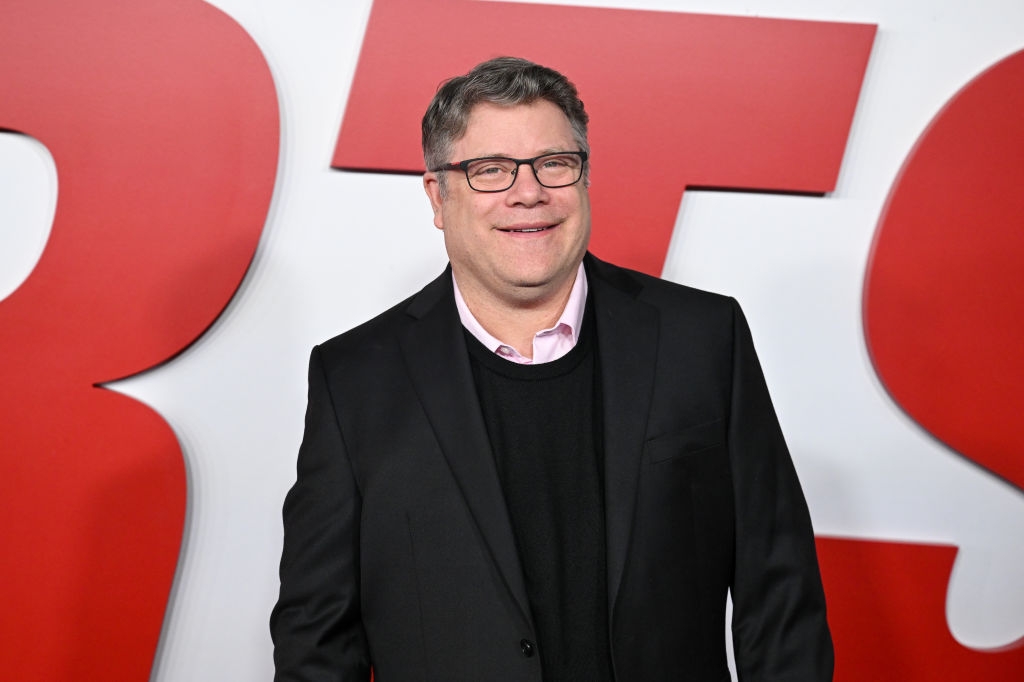 Person in a black suit and sweater over a shirt on a red carpet, smiling in front of large stylized letters