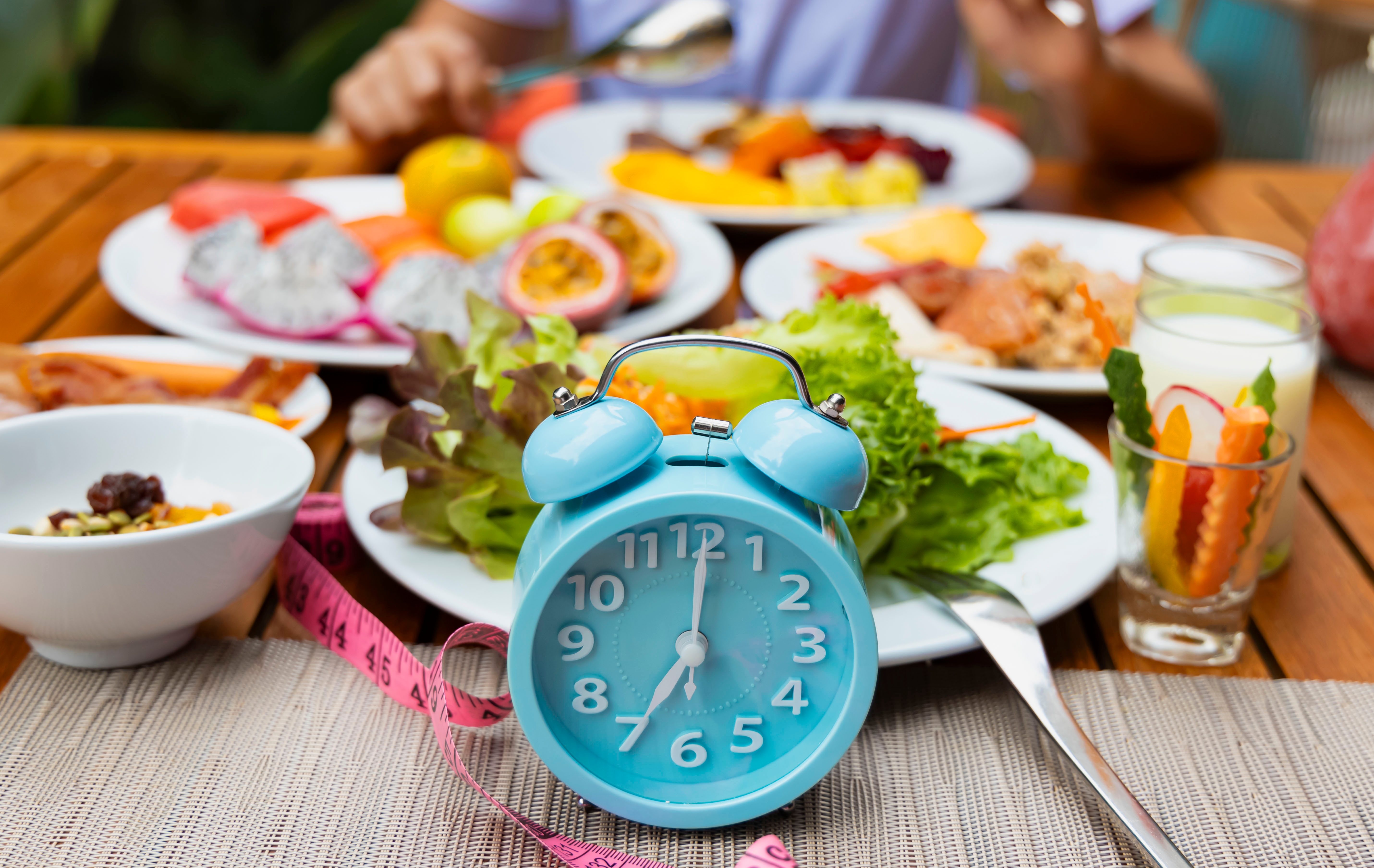 A blue alarm clock sits on a table with a variety of dishes, including fruits and salads, suggesting a focus on meal timing or diet