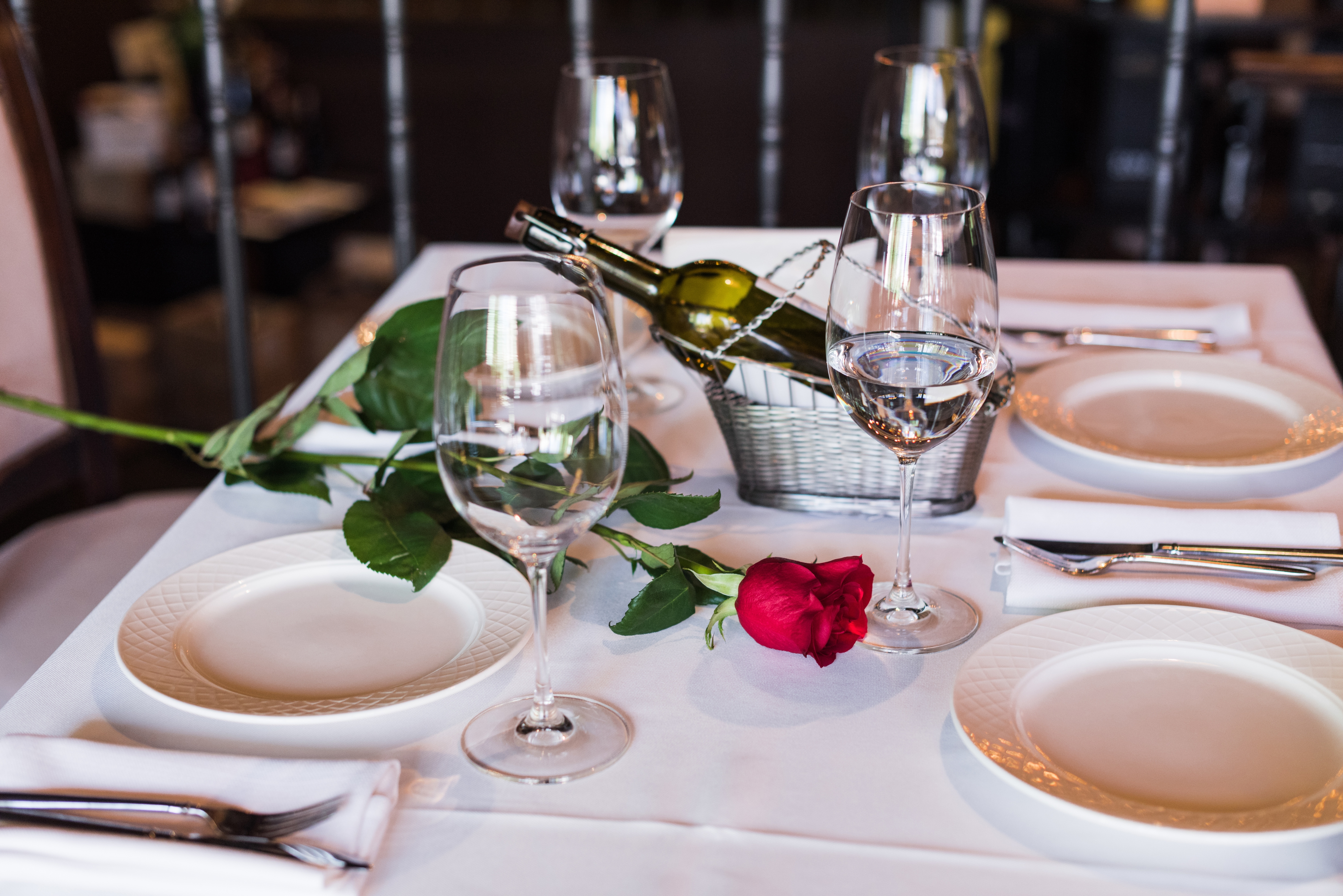 Elegant table setting with wine, glasses, plates, and a single red rose on a white tablecloth, suggesting a romantic dinner