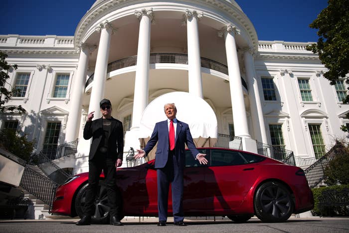 Elon Musk and Donald Trump stand gesturing in front of the White House, alongside a Tesla on display