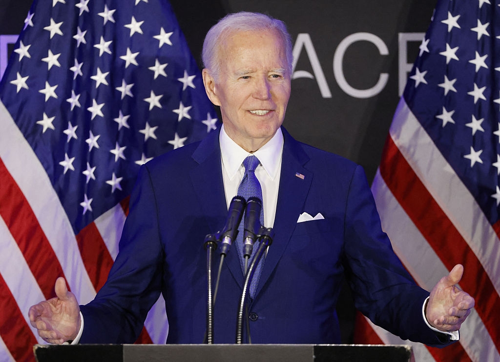 Joe Biden stands at a podium with microphones, American flags in the background, addressing an audience