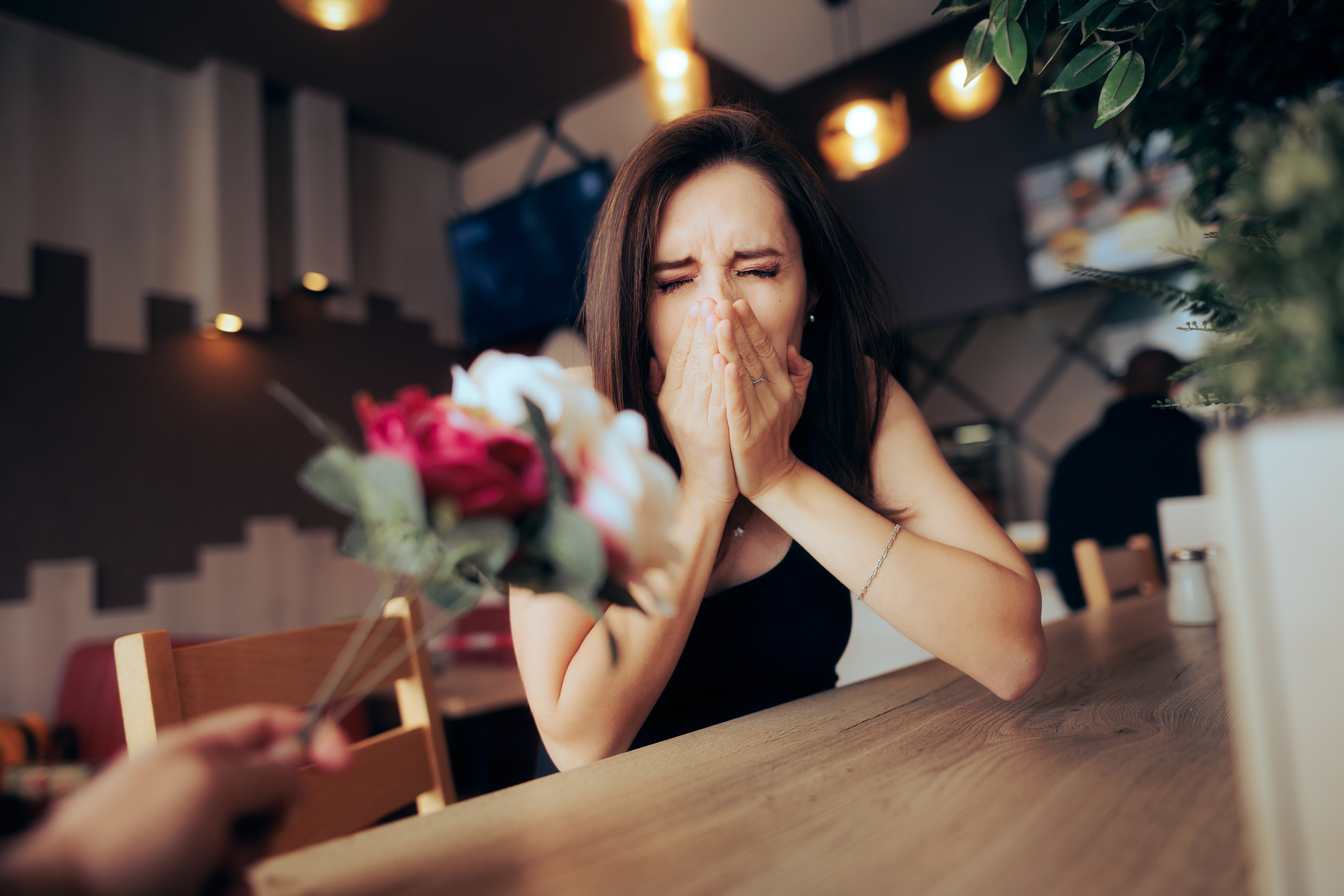 A person is sitting at a table, reacting with surprise and delight as someone presents them with a bouquet of flowers
