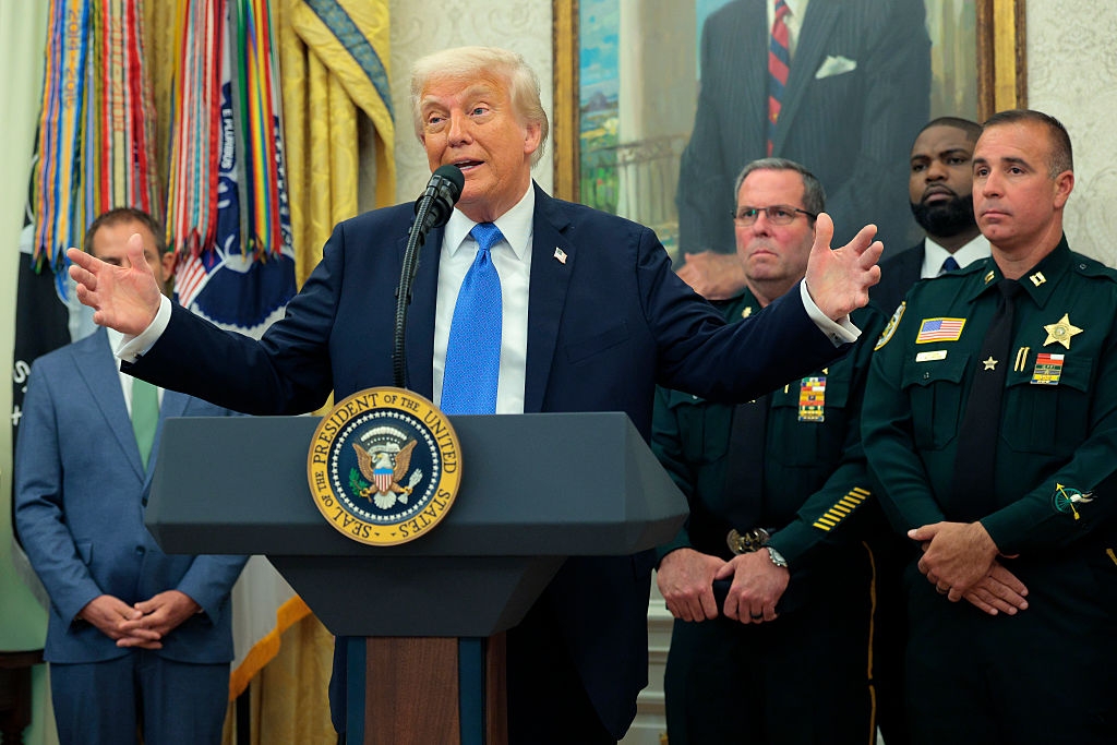 Donald Trump speaks at a podium with officials standing behind in a formal setting, including law enforcement in uniform