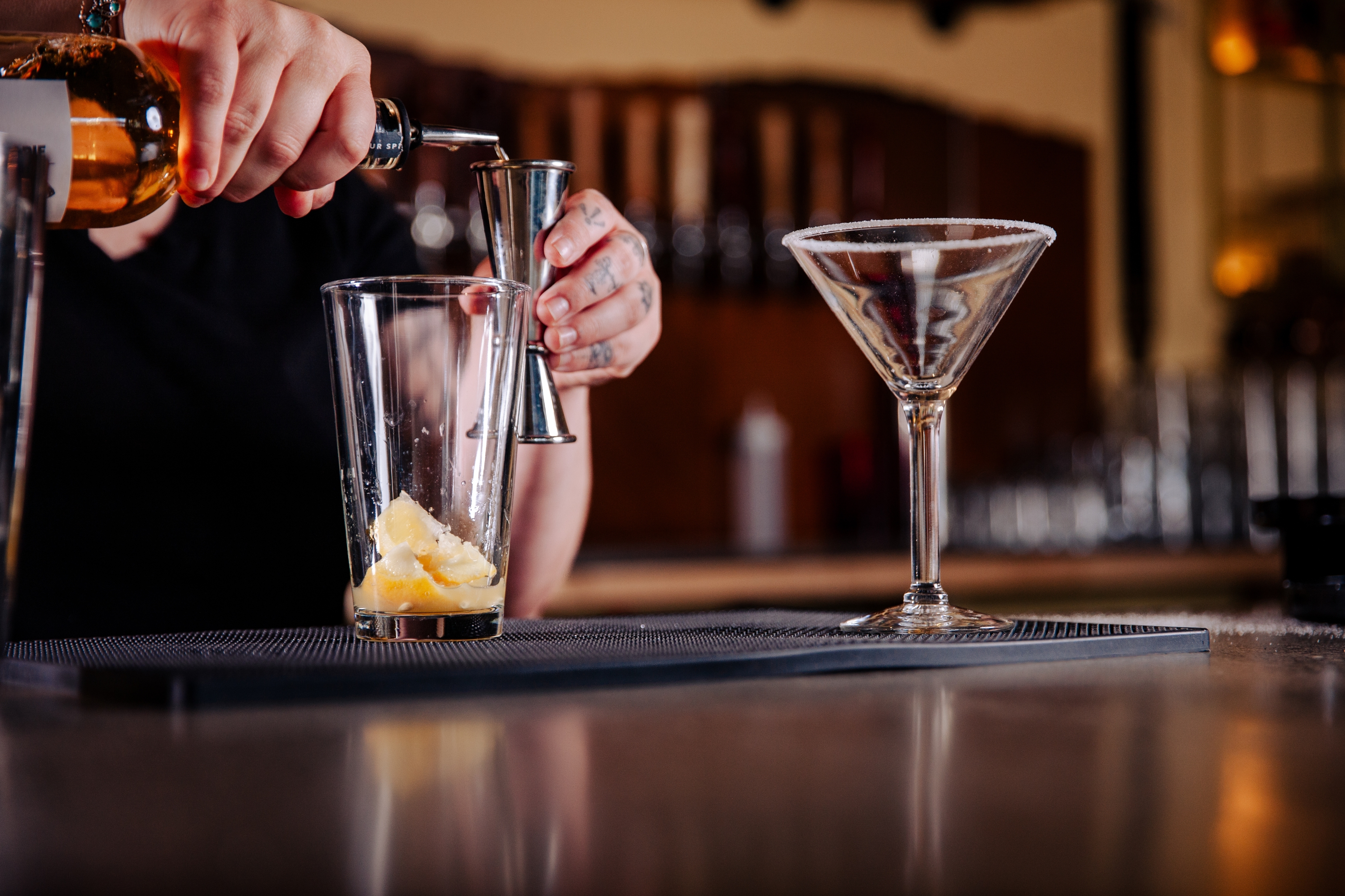 Person preparing a cocktail, measuring and pouring drink ingredients into a glass, with a ready martini glass in the foreground